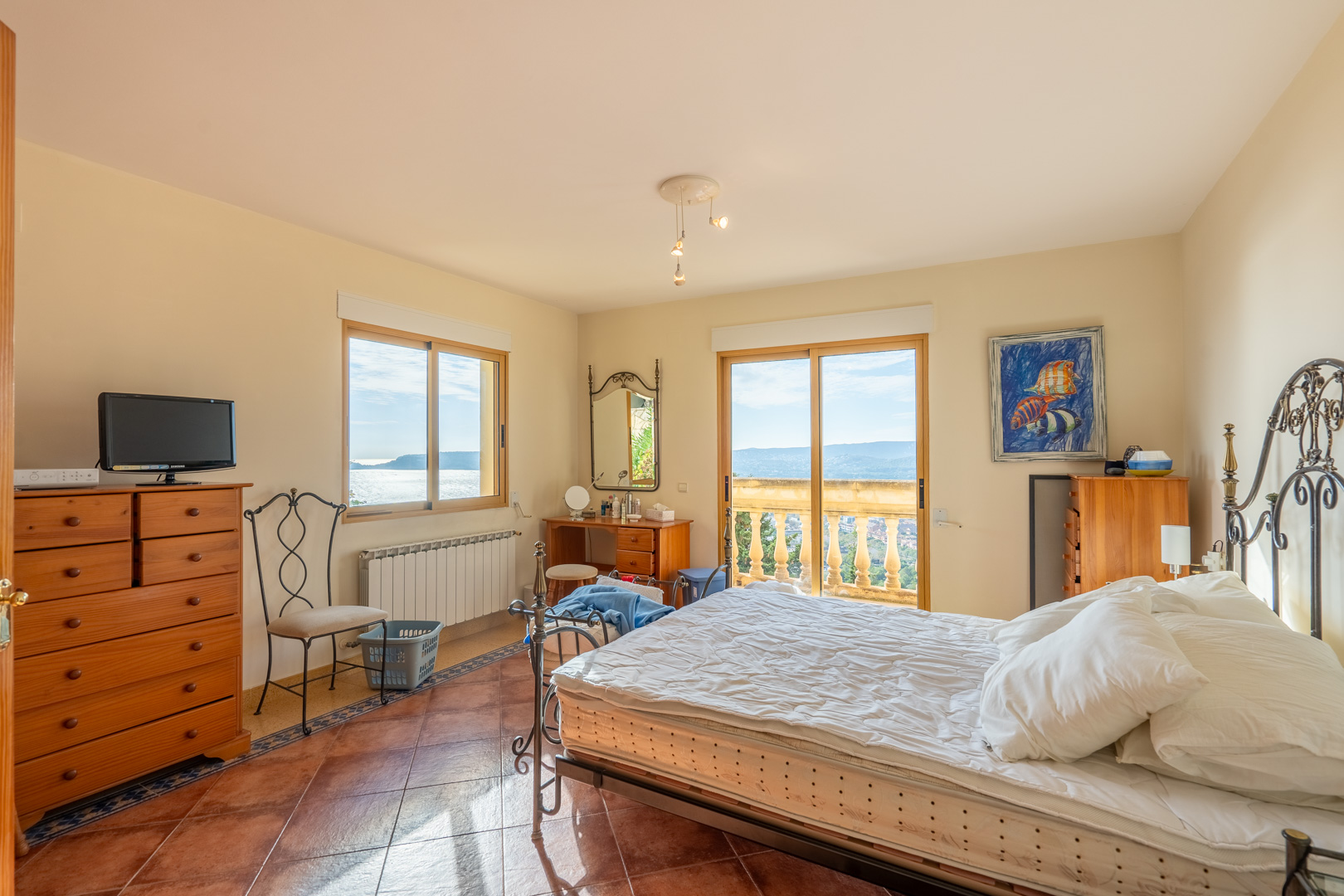 Bedroom featuring terracotta tile flooring, a wrought iron bed, and sliding doors opening to a balcony with stone balustrades and expansive Mediterranean views.