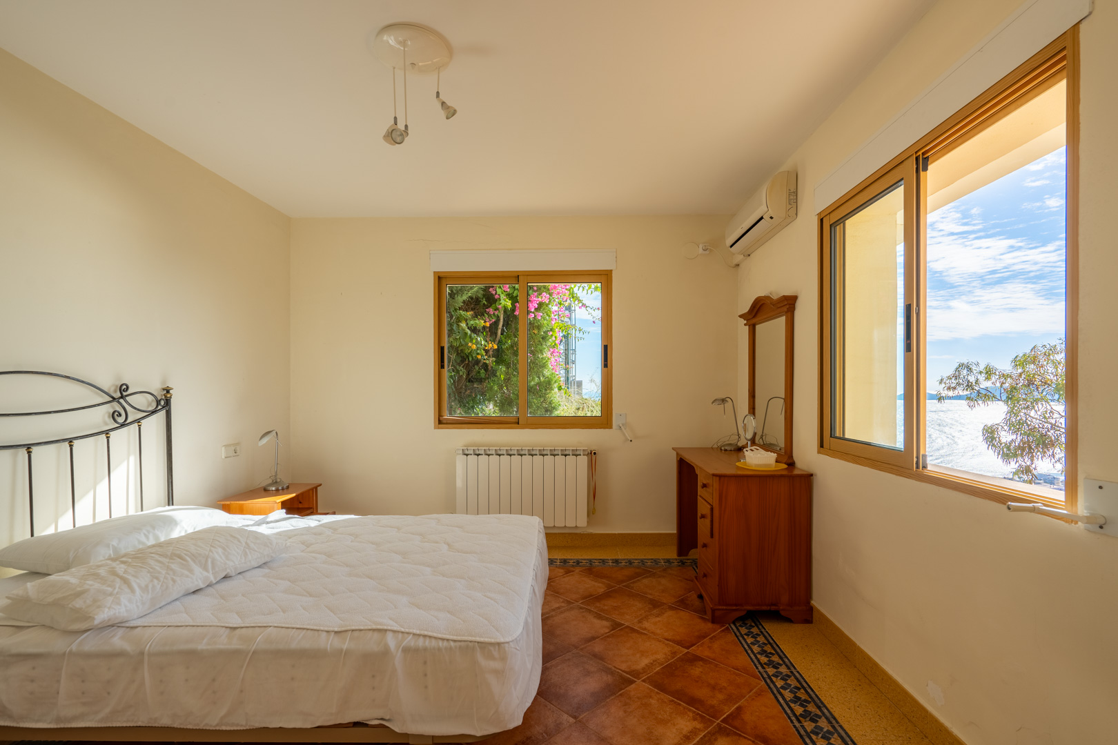 Bedroom featuring terracotta tile flooring with decorative borders. Includes wooden-framed windows with sea views, a wrought iron headboard, and split air conditioning.