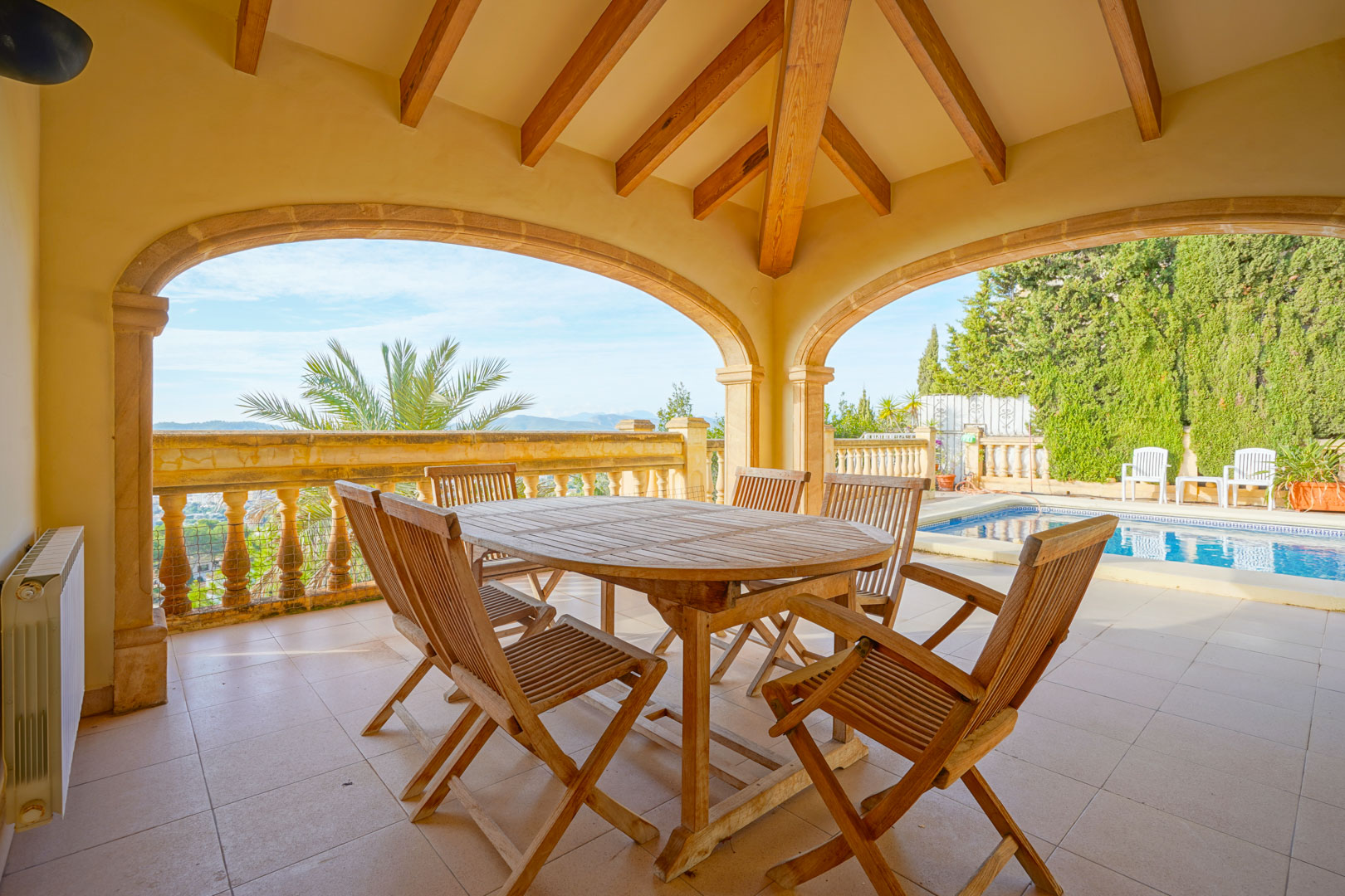 Covered terrace featuring exposed wooden beams, stone arches, and a wooden dining set overlooking the private pool and mountain landscape.