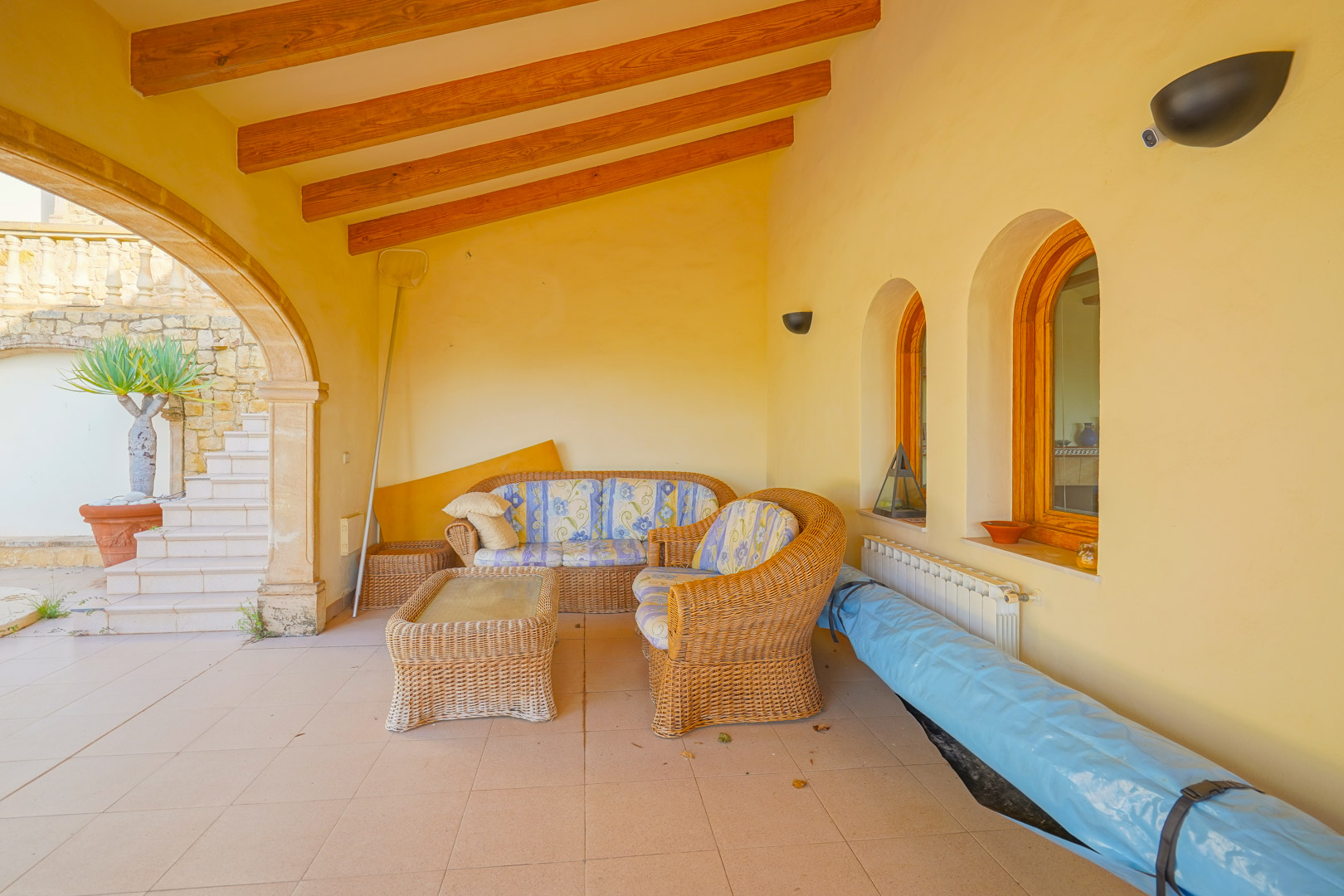 Covered terrace featuring exposed wooden beams, traditional arched windows, and a wicker lounge set on a tiled floor adjacent to the pool area.