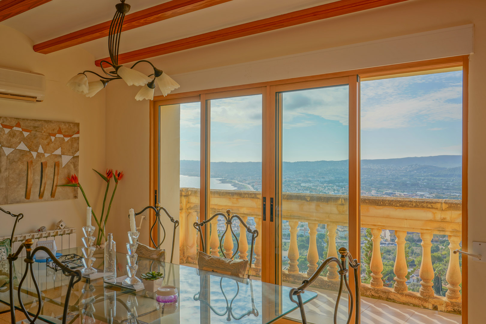 Dining area featuring a glass table and wooden ceiling beams, with large sliding doors overlooking the Jávea coastline and stone terrace balustrade.