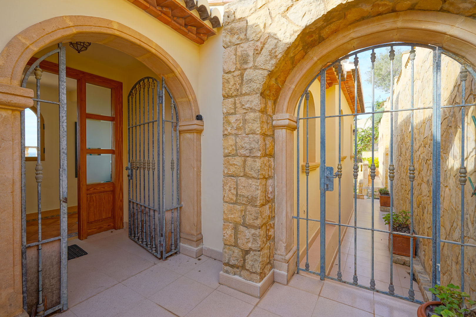 Entrance area featuring traditional Tosca stone arches, decorative wrought iron gates, and a wooden main door with glass panels.