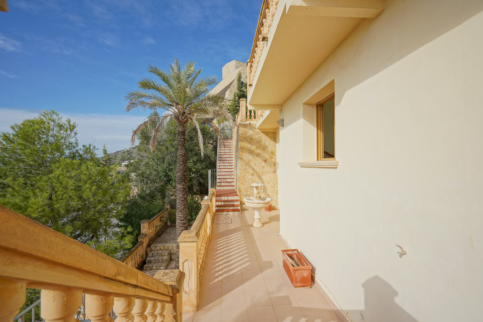 Exterior walkway featuring classic balustrades, natural stone walls, and stairs with traditional decorative tiles set against Mediterranean greenery.