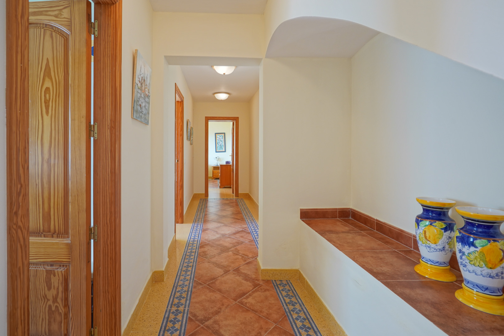 Hallway featuring terracotta-style floor tiles with decorative borders, natural wood doors, and a built-in tiled bench with traditional ceramic vases.