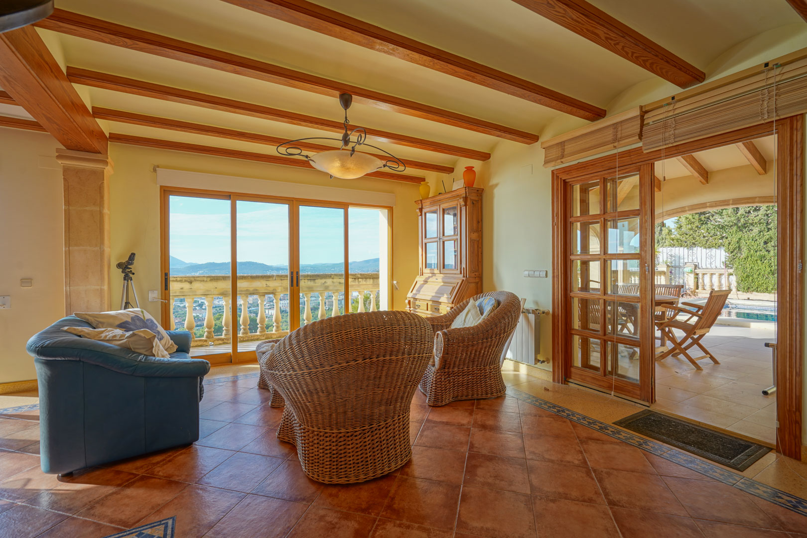 Living space featuring exposed wooden beams, terracotta tiles, and wicker armchairs. Large glass doors frame panoramic valley views and lead to the poolside terrace.