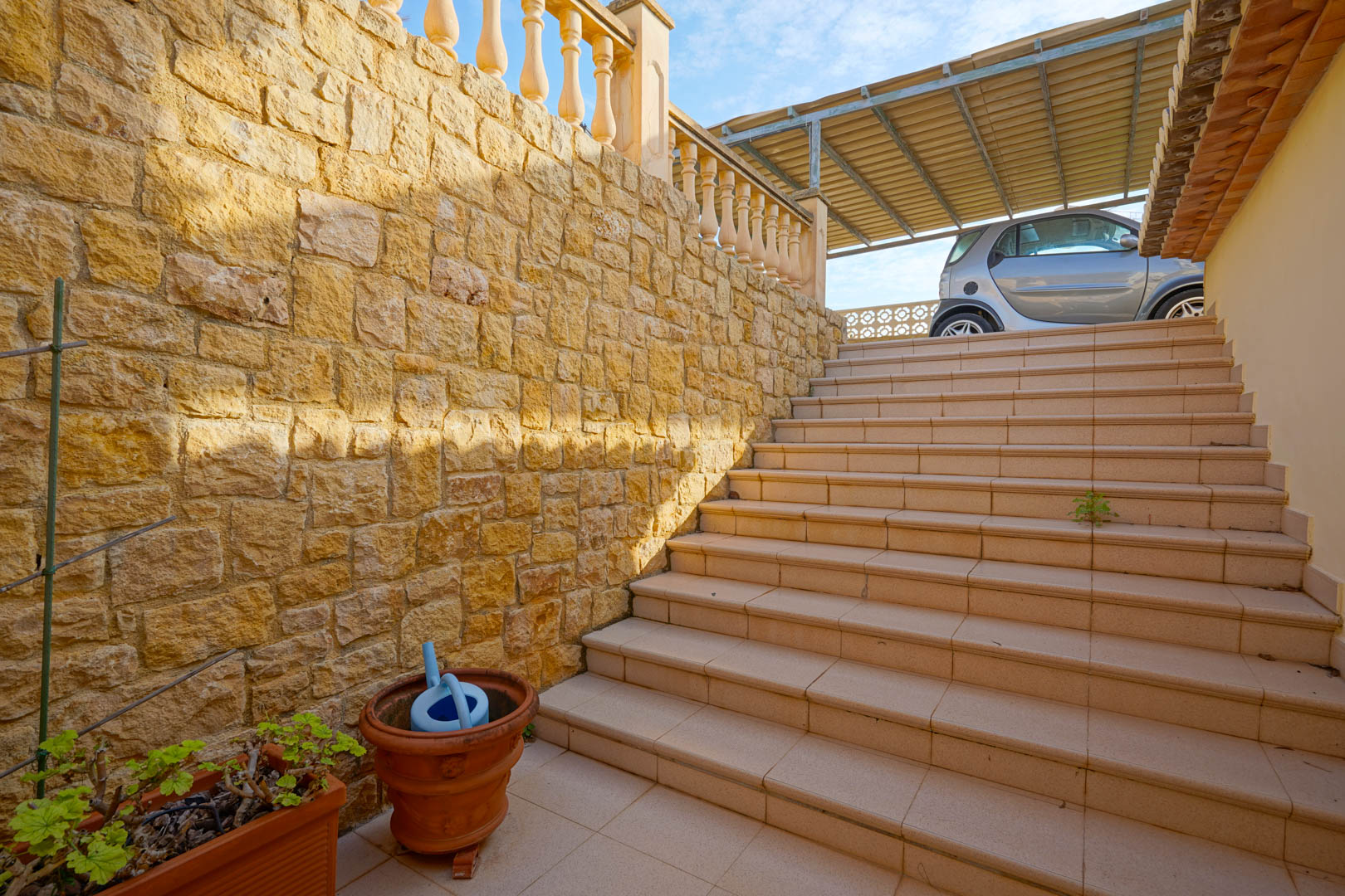 Outdoor staircase bordered by a textured natural stone wall leading to the upper parking level and a covered carport for vehicles.
