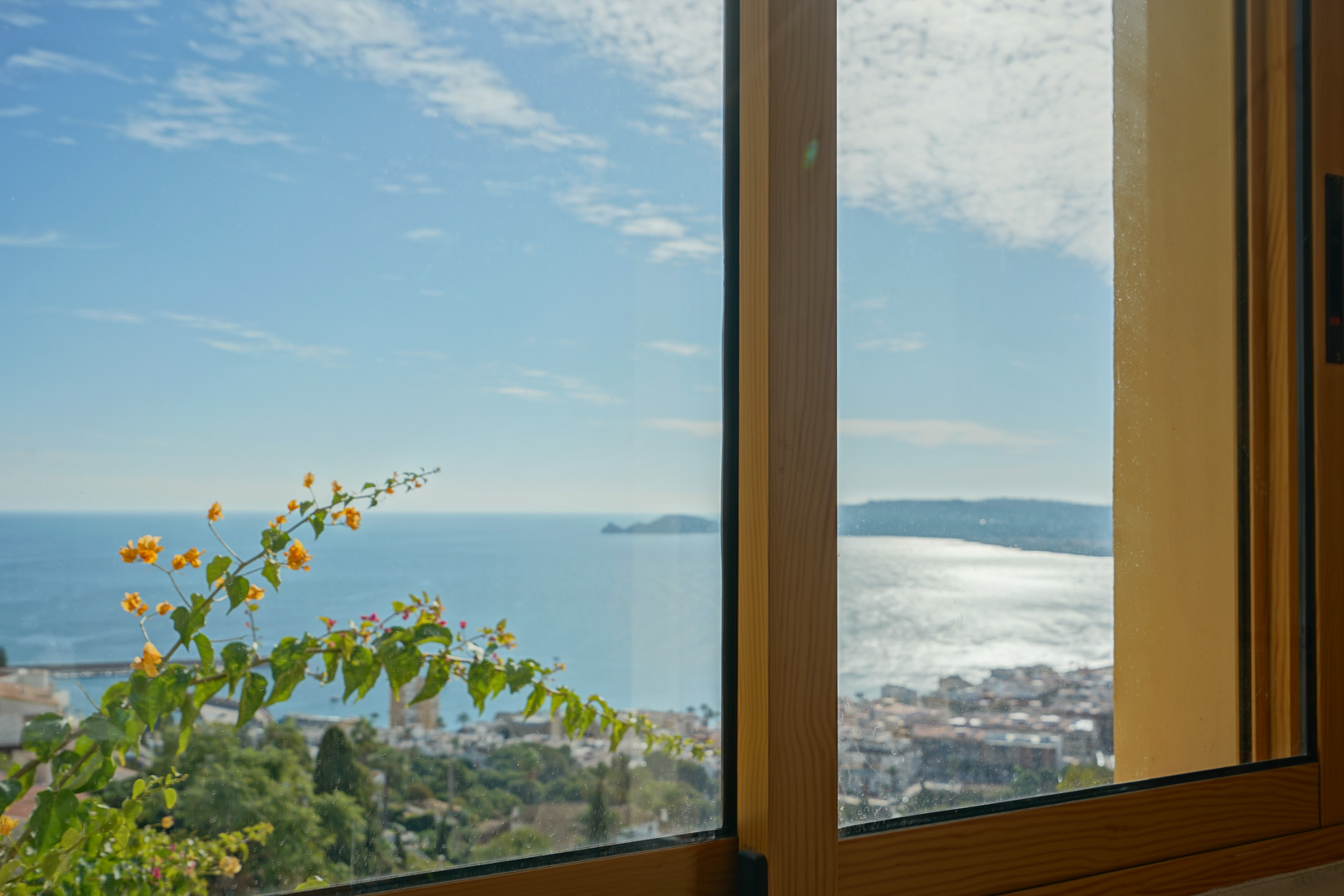 Panoramic view of Jávea Bay and Cap Negre through a wooden-framed window, framed by vibrant flowering bougainvillea.