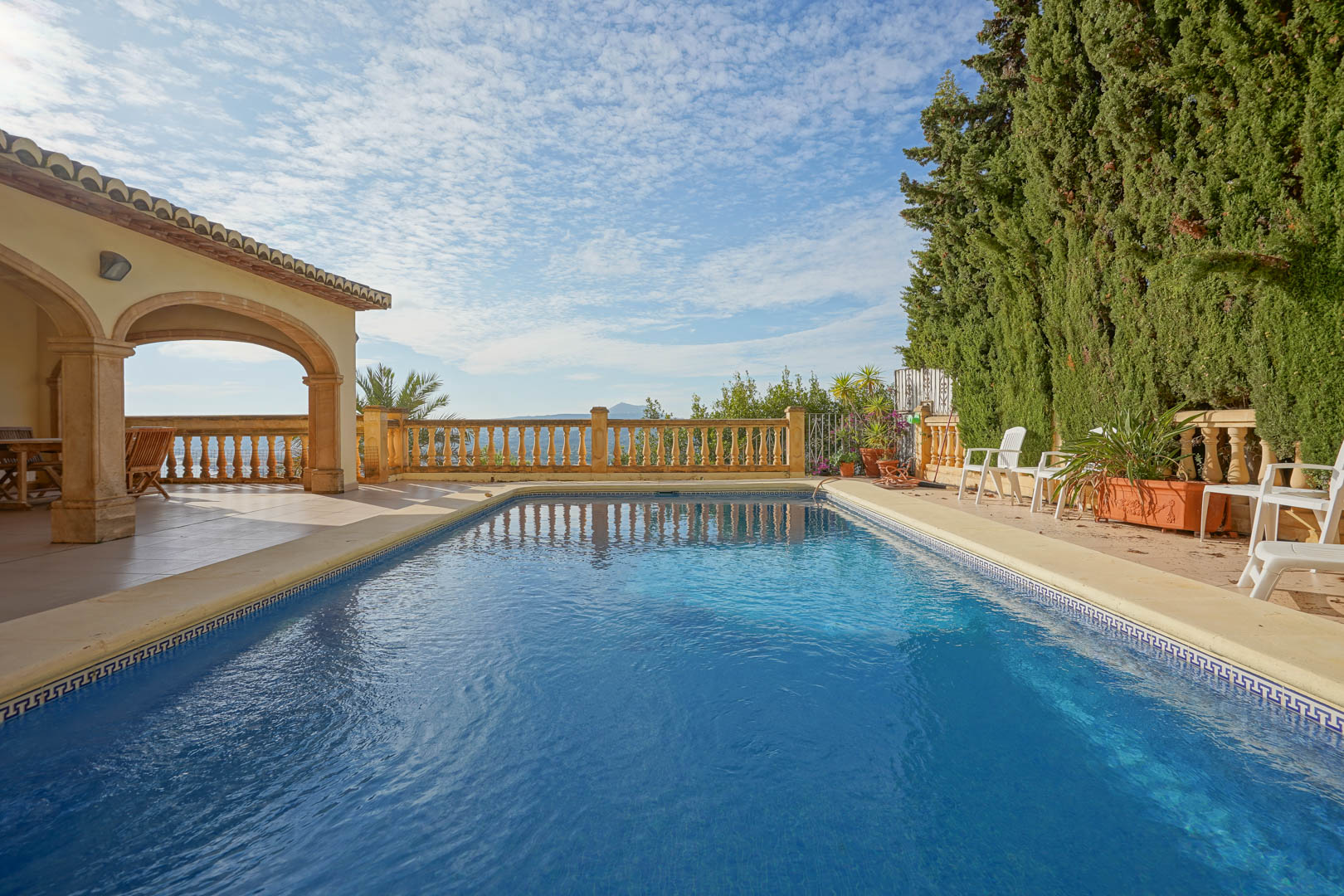 Rectangular swimming pool featuring a stone border, adjacent to a traditional arched porch and a classic balustrade overlooking the Mediterranean horizon.