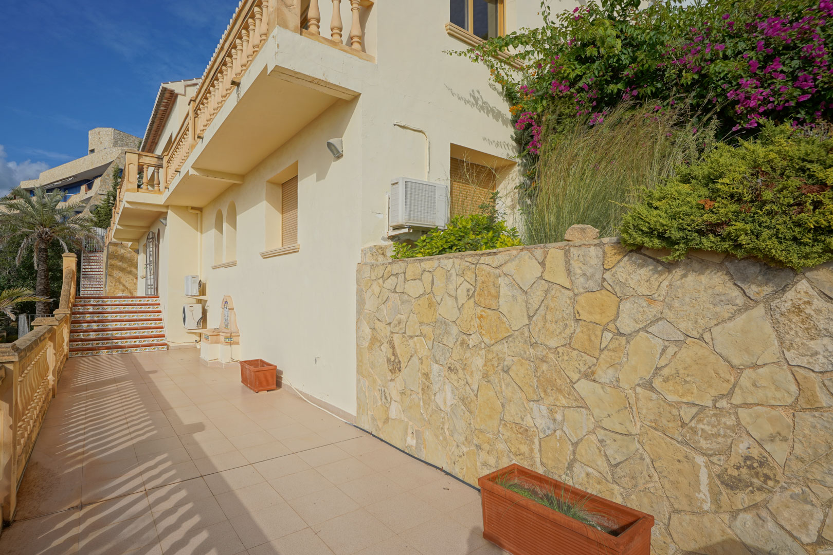 Side terrace featuring a natural stone wall, terracotta planters, and stairs with traditional decorative tiles. The facade shows classic Mediterranean architectural details.