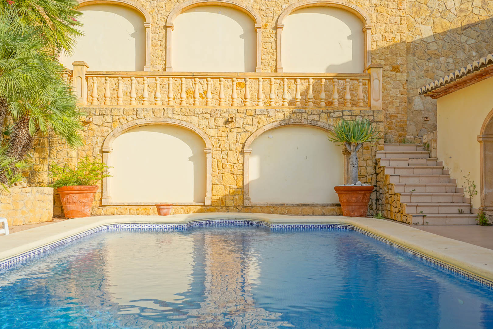 Swimming pool featuring blue mosaic tiling and a Greek key border, set against a natural stone wall with arched niches, a classic balustrade, and terracotta planters.
