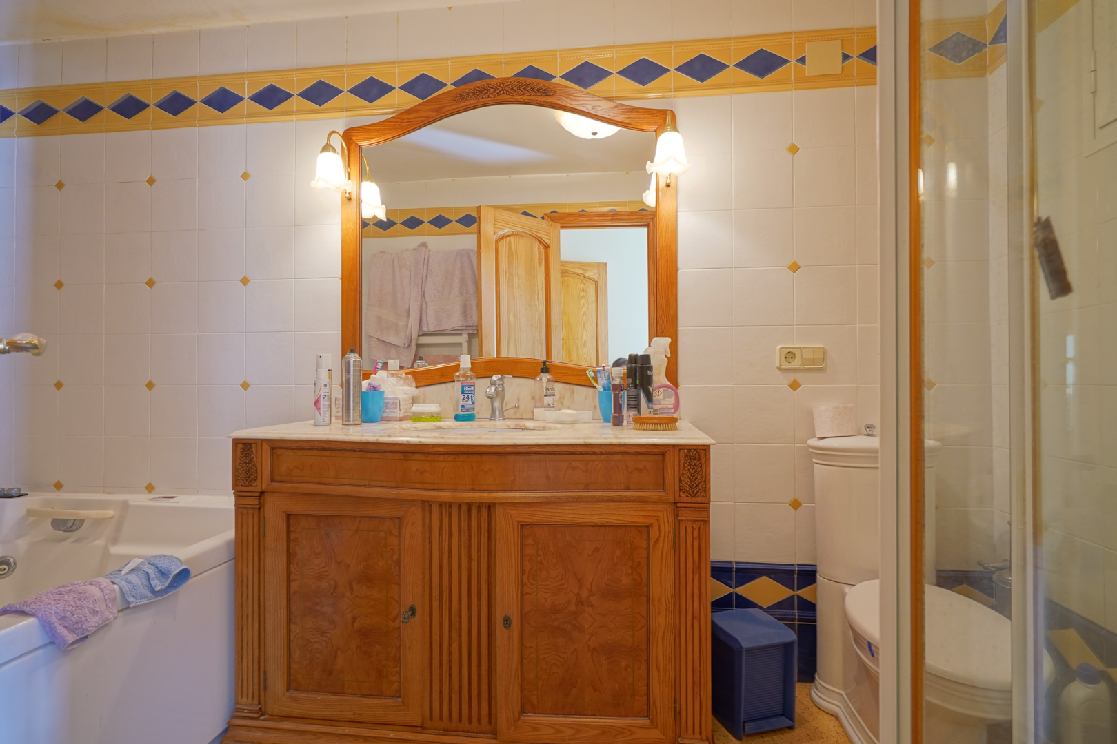 Traditional bathroom featuring a carved wooden vanity with a marble top, framed mirror with classic wall lamps, and decorative tiled walls.