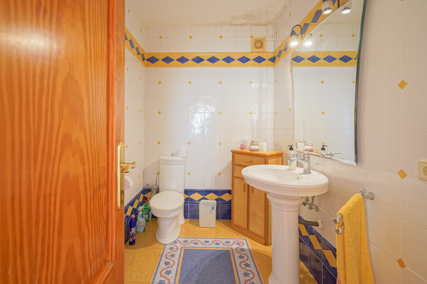 Traditional bathroom featuring white wall tiles with blue and yellow diamond accents, a classic pedestal sink, and a wooden storage cabinet.