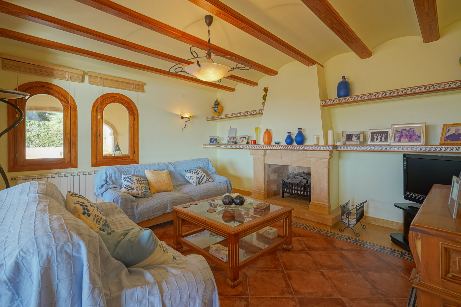 Traditional living room featuring a stone fireplace, exposed wooden ceiling beams, and terracotta tile flooring. Includes arched wooden windows and built-in shelving.