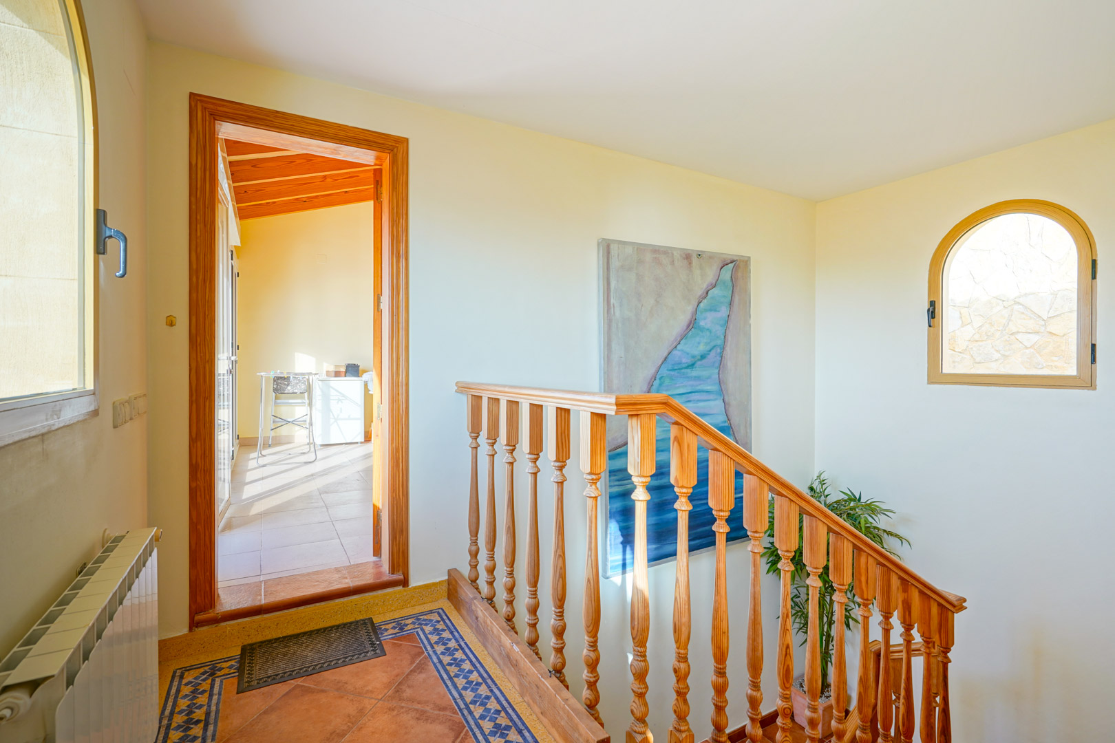 Upper-level landing featuring a traditional wooden staircase, decorative tiled flooring, and an arched window. An open doorway reveals a room with a sloped wooden ceiling.
