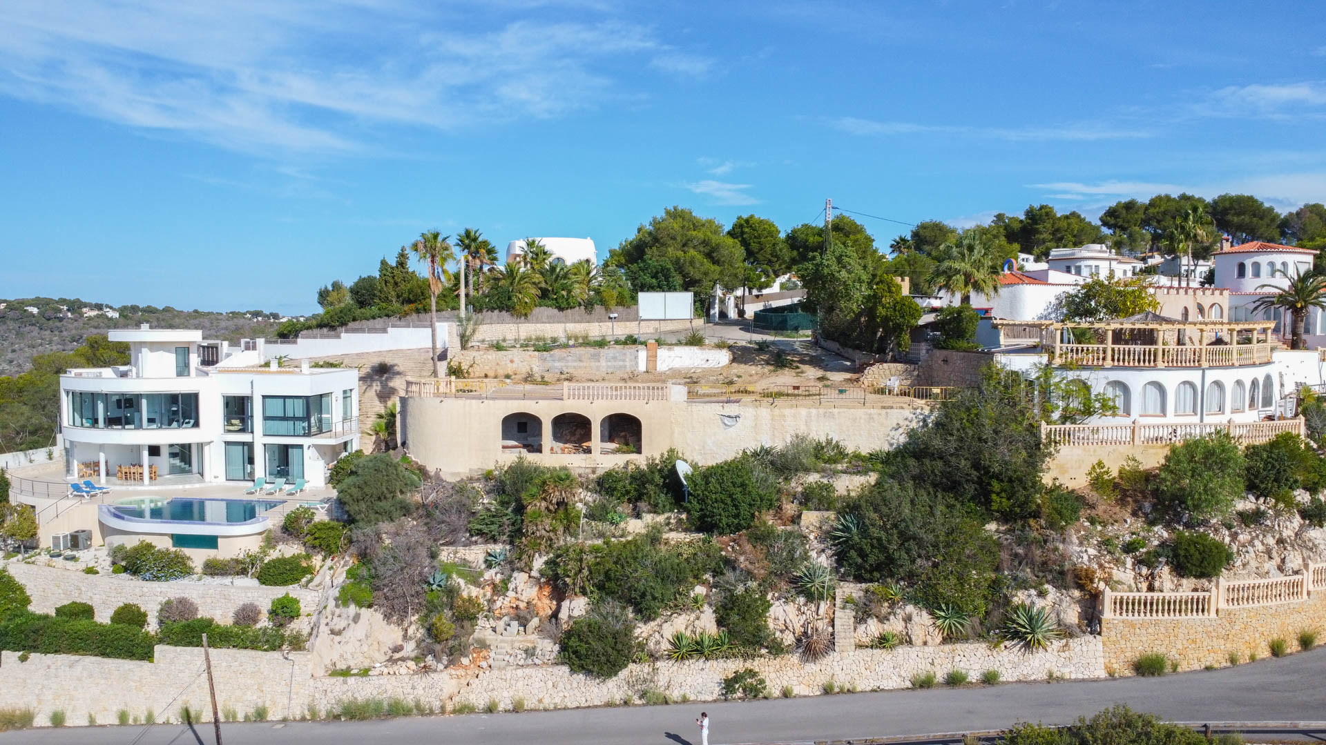 Aerial view of the elevated plot featuring an arched stone foundation, situated between luxury villas with native Mediterranean landscaping and stone retaining walls.