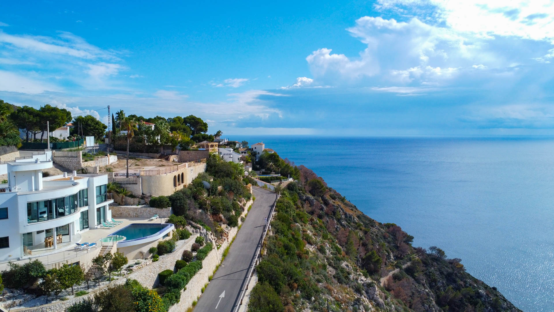 Aerial view of the elevated plot featuring panoramic Mediterranean Sea views, adjacent to modern villas and a winding coastal access road.