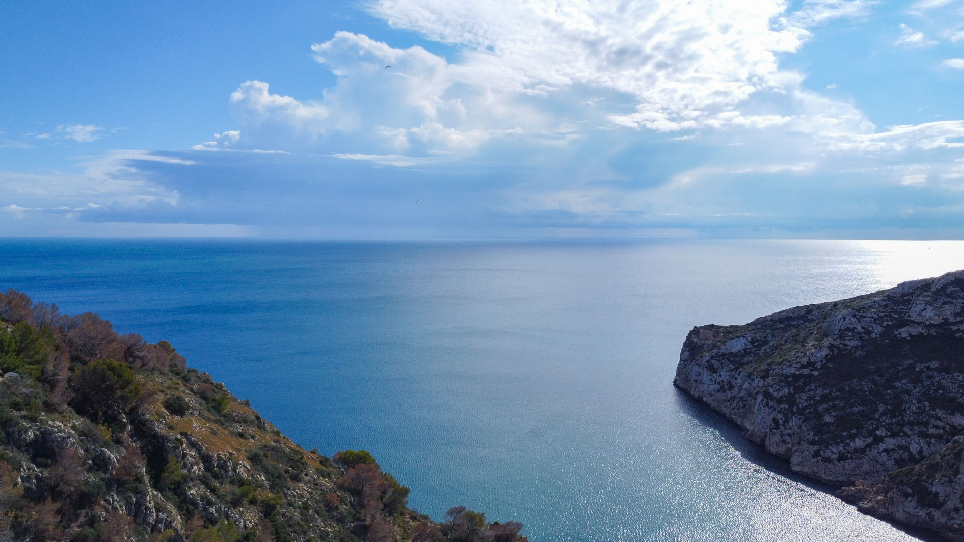Frontal aerial view of the Mediterranean Sea and the rocky cliffs flanking the Granadella bay under a partially cloudy sky.