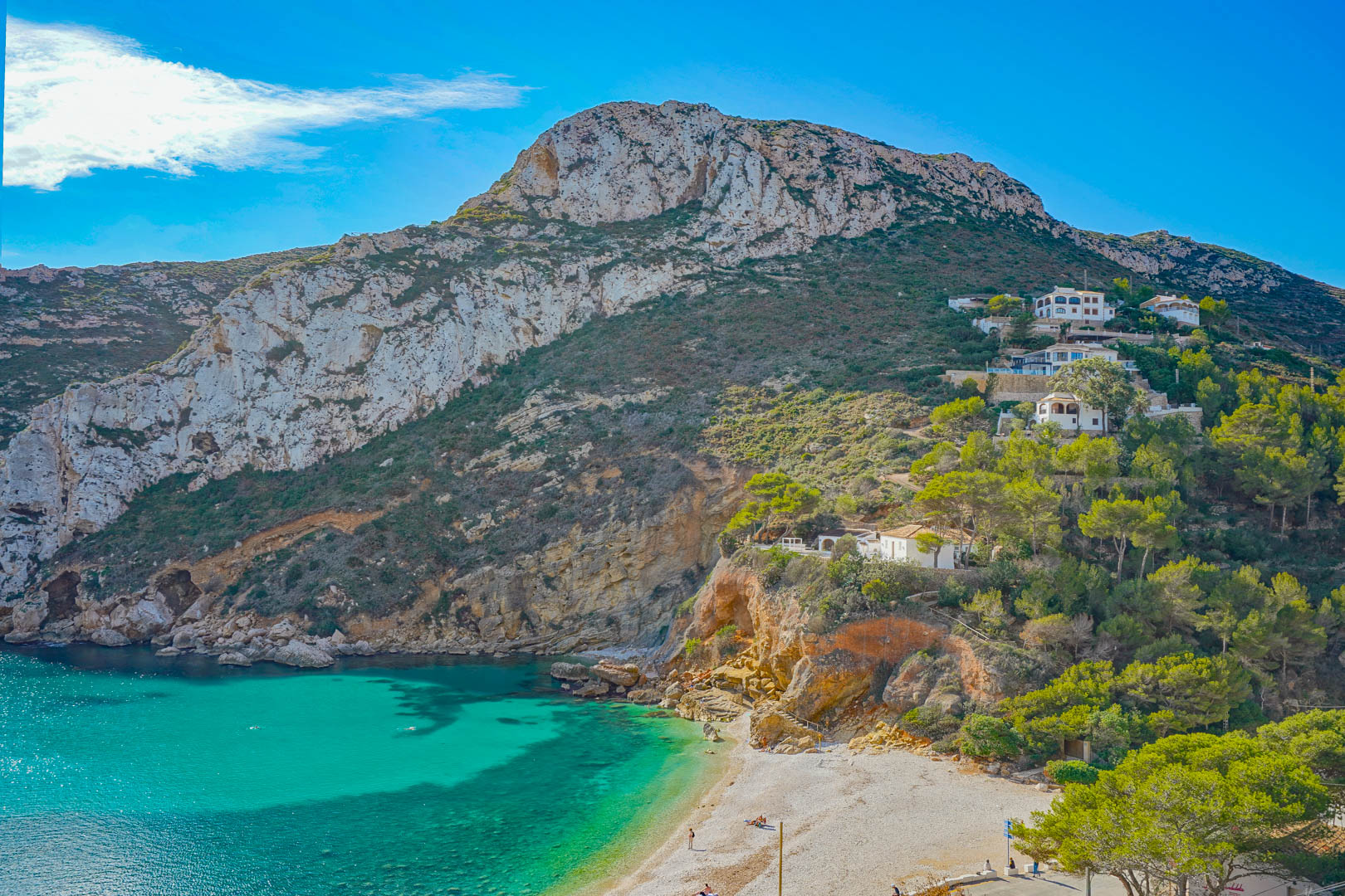 Panoramic view of Granadella Cove featuring turquoise waters, limestone cliffs, and Mediterranean pine forests surrounding the pebble beach.