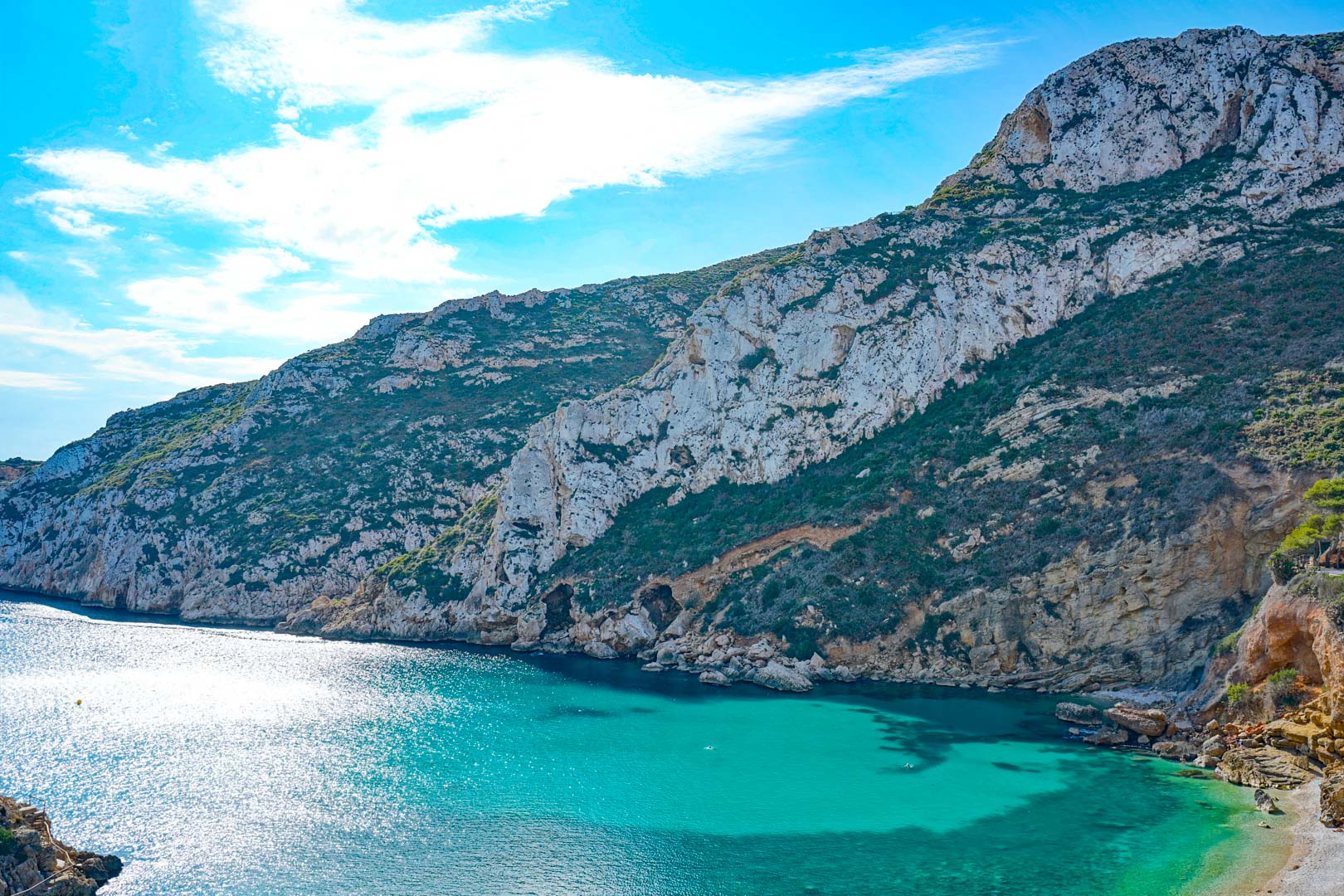 Panoramic view of the turquoise waters at Granadella Bay, framed by rugged limestone cliffs and Mediterranean shrubland under a bright sky.