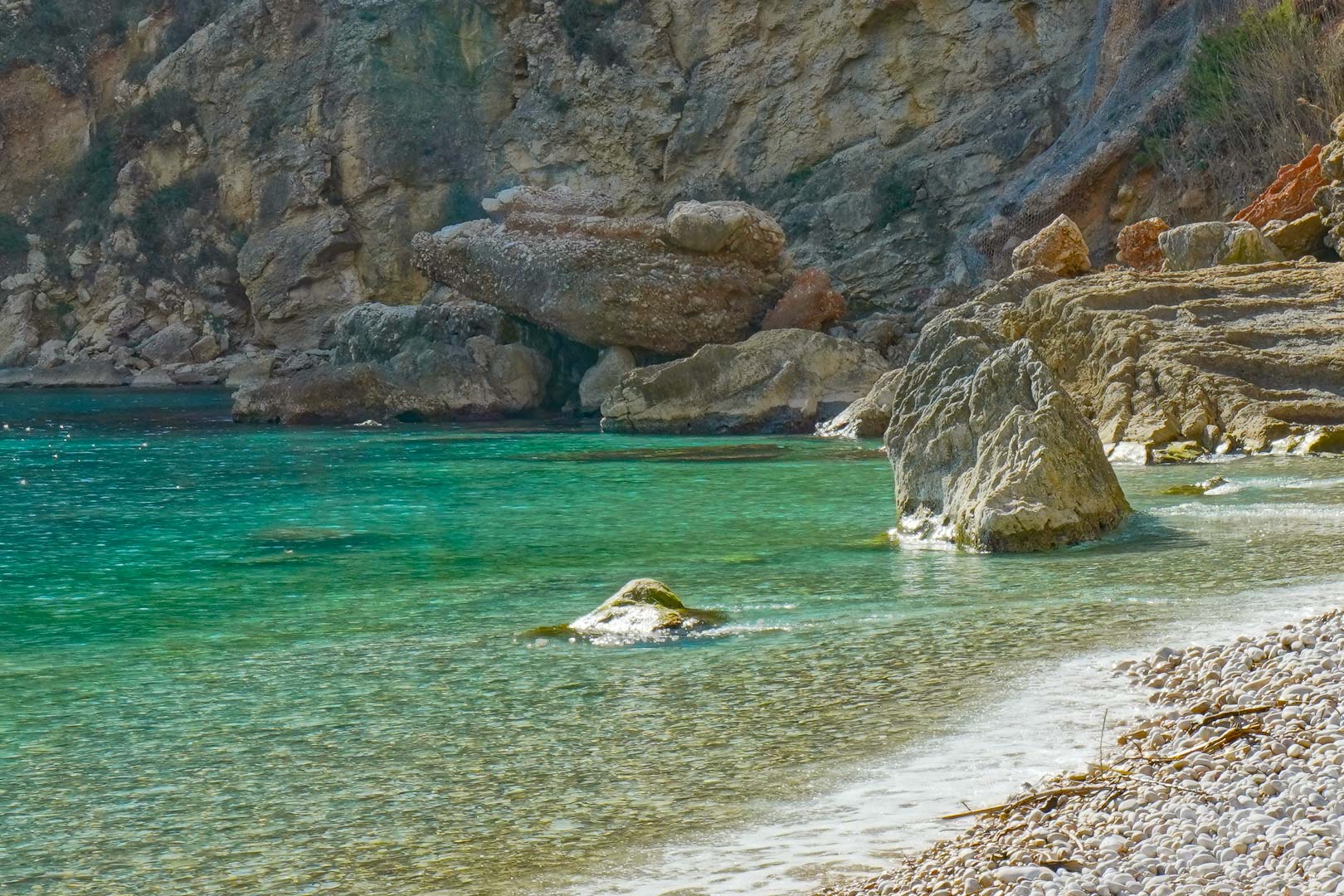 View of the pebble shoreline at Cala Granadella, featuring crystal-clear turquoise waters and the surrounding natural limestone rock formations.