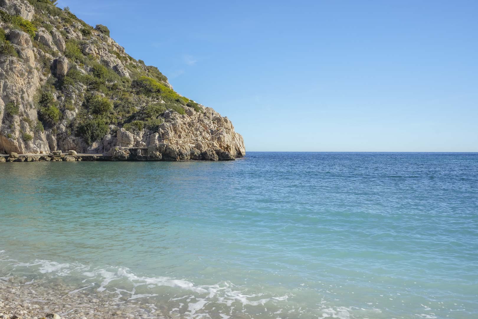 View of the turquoise waters at Granadella Bay, featuring a pebble shoreline and rugged limestone cliffs with native Mediterranean vegetation.