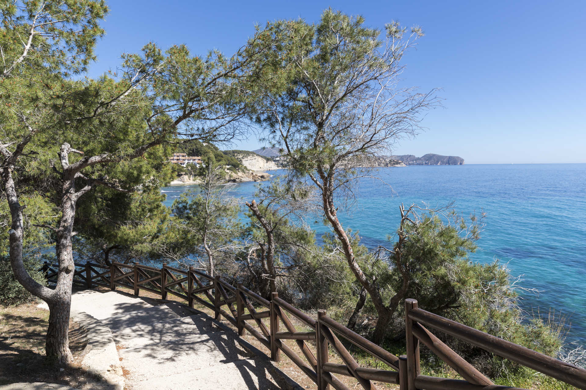 Coastal walking path featuring a wooden railing and pine trees, overlooking the turquoise Mediterranean waters near La Fustera beach.