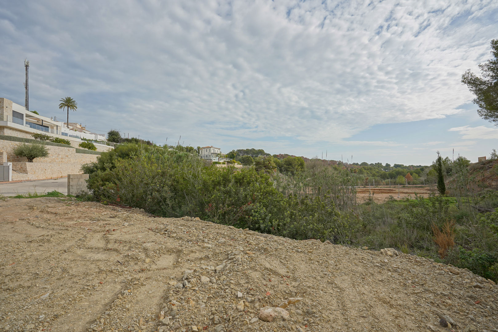 Construction-ready plot featuring cleared terrain surrounded by Mediterranean greenery and established modern villas with natural stone walls.