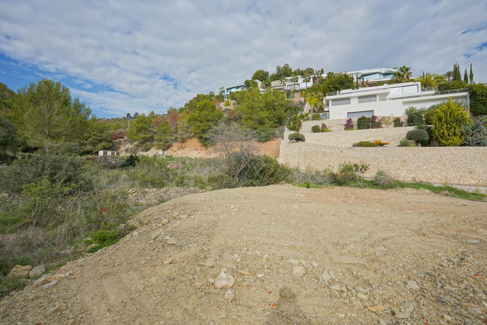 Elevated building plot under development, featuring views of neighboring modern white villas and tiered dry stone retaining walls set against a pine-covered hillside.