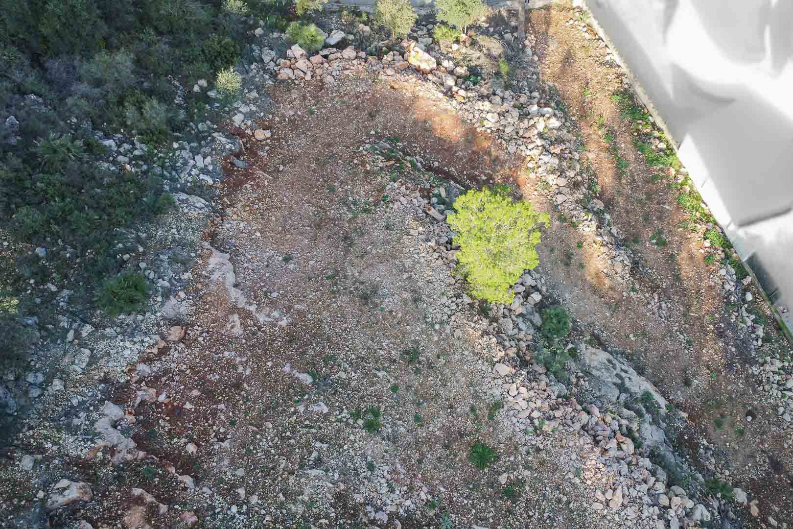 Aerial view of the building plot featuring rocky terrain, native shrubs, and a young pine tree. The land is prepared for construction with a clear perimeter.