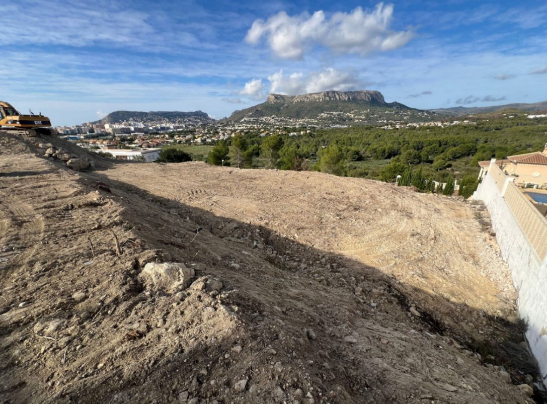 Elevated construction site showing initial earthworks with panoramic views of the Peñón de Ifach, Calpe town, and the Mediterranean coastline.