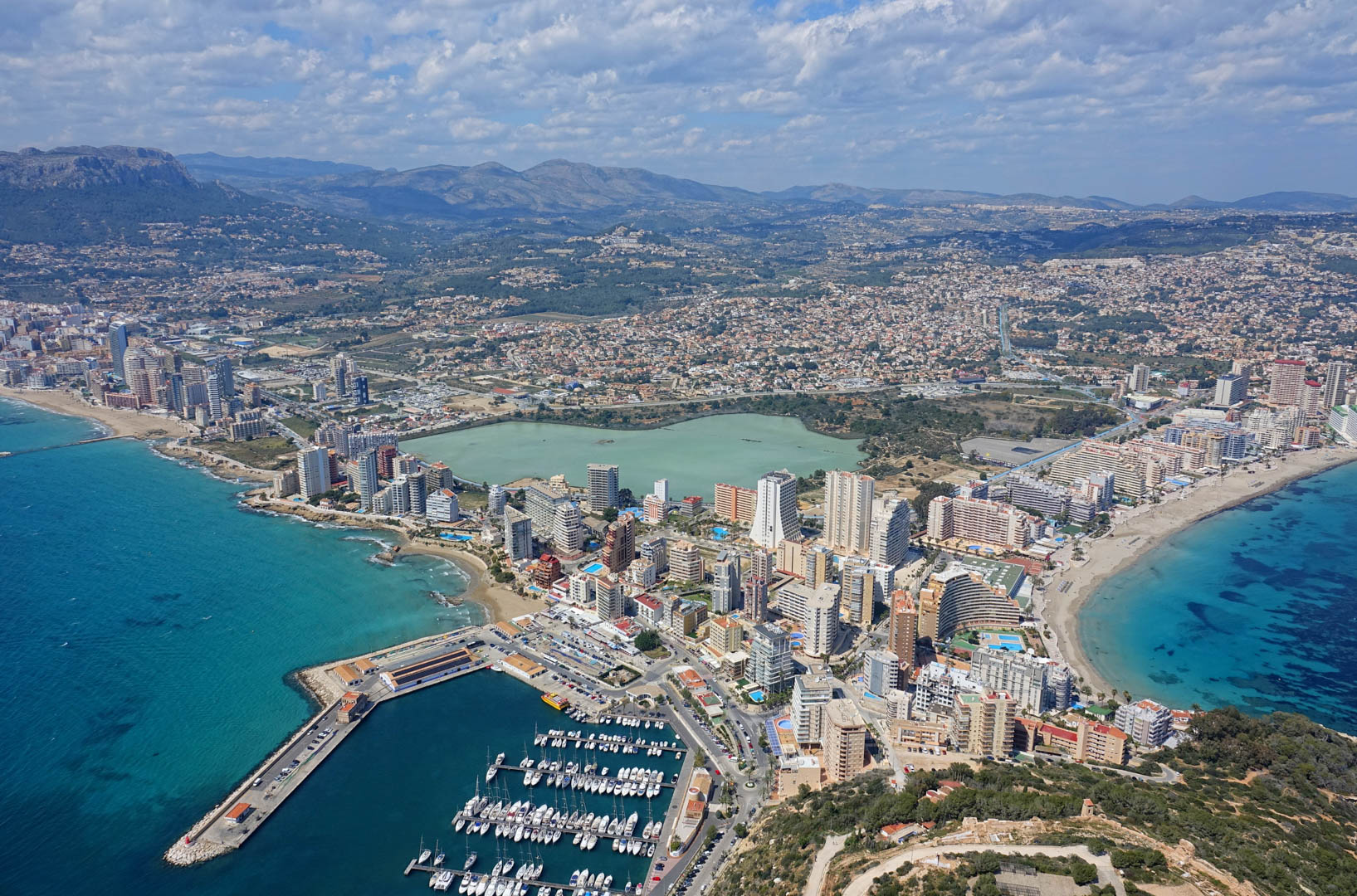 Panoramic aerial view of Calpe featuring the marina, sandy beaches, Las Salinas salt lake, and the urban skyline set against distant mountain ranges.