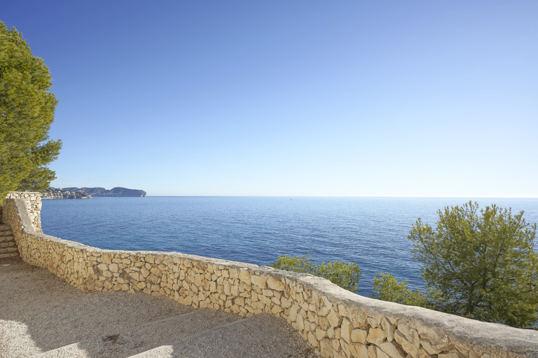 Panoramic Mediterranean Sea views from a terrace featuring a traditional dry stone wall. The scene includes native pine trees and a clear horizon over the blue water.
