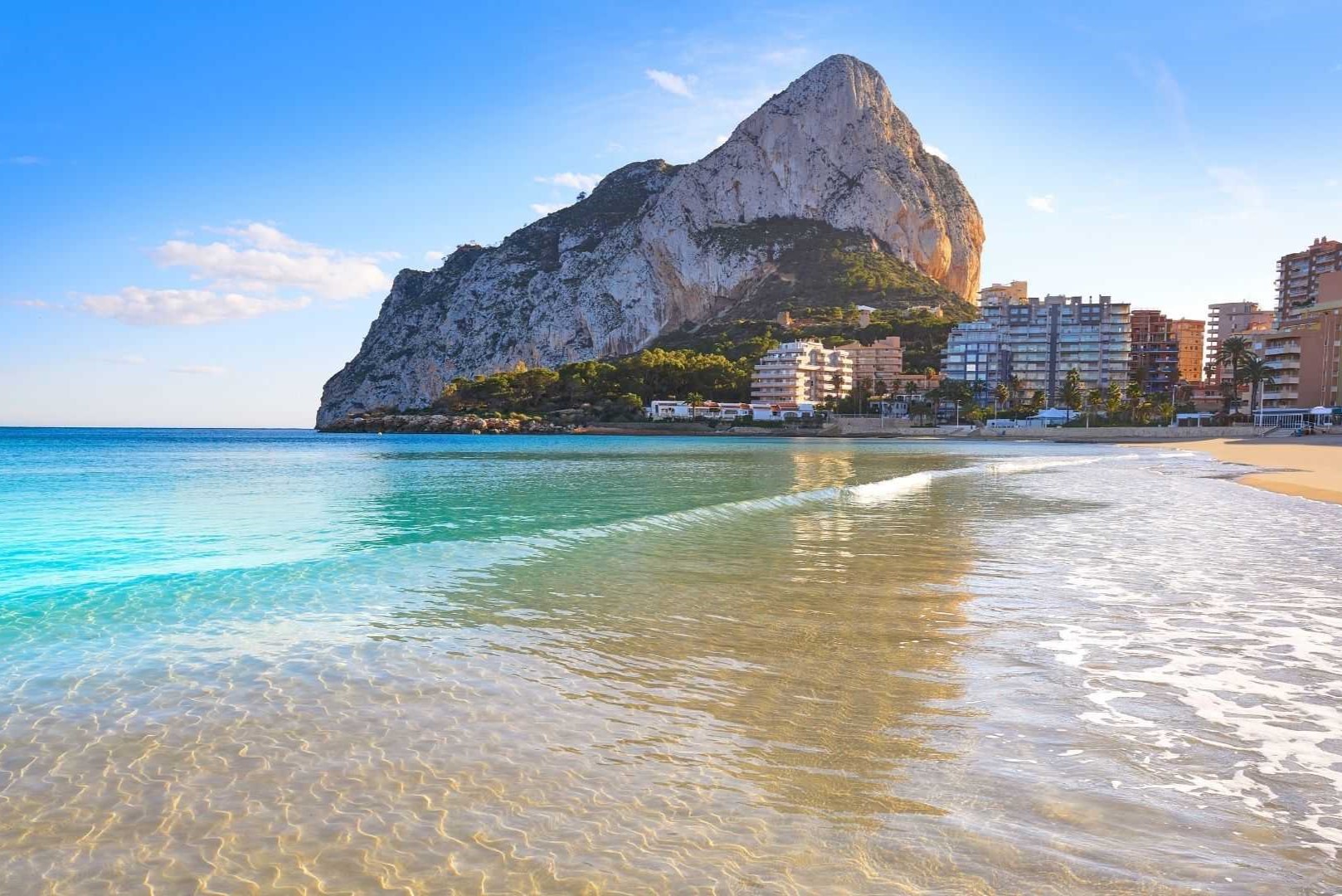 Panoramic view of the Peñón de Ifach from the Calpe shoreline, featuring crystal-clear Mediterranean waters and the coastal skyline under a bright, clear sky.
