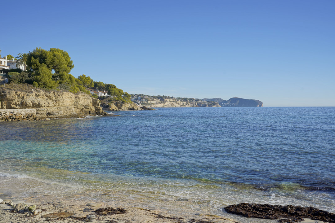 View of a Calpe cove featuring turquoise waters, limestone cliffs, and Mediterranean pine trees under a clear blue sky.