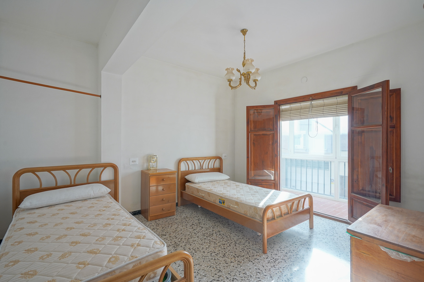 Bedroom featuring twin beds, original terrazzo flooring, and wooden doors opening onto a private balcony with natural light.