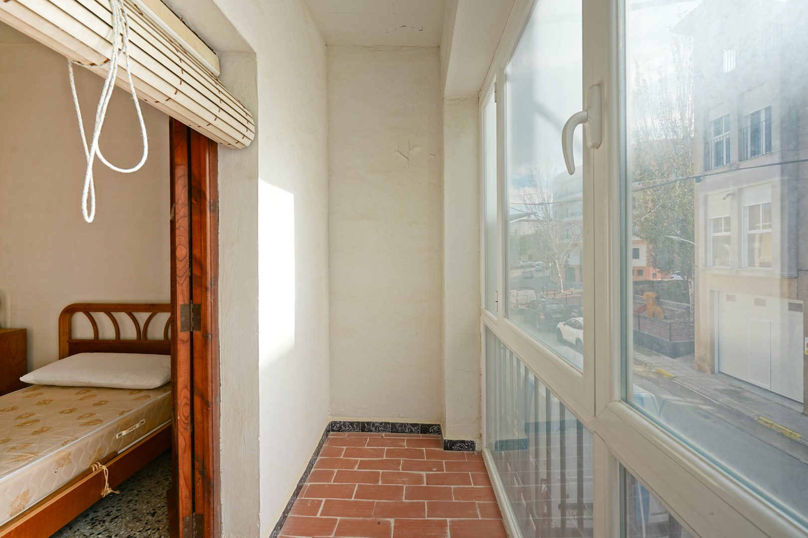 Enclosed sunroom featuring terracotta tile flooring, white PVC window frames with street views, and direct access to an adjacent bedroom through wooden doors.