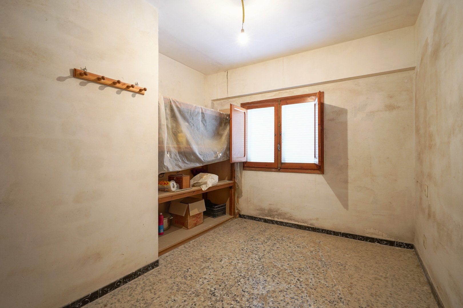 Room featuring traditional terrazzo flooring, a double-leaf wooden window, and built-in shelving, currently in need of complete renovation.