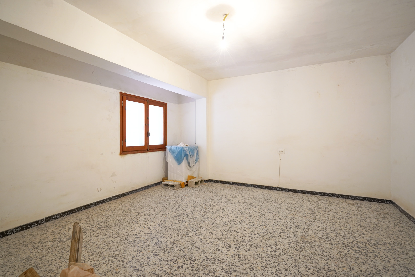 Spacious room featuring original terrazzo flooring, white walls awaiting renovation, and a wooden-framed window providing natural light.