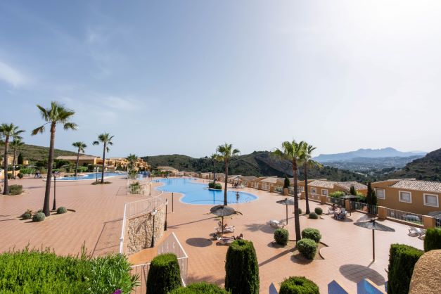 Communal area of Monte Cala Gardens featuring two large swimming pools, a spacious sun terrace with palm trees, and panoramic mountain views.