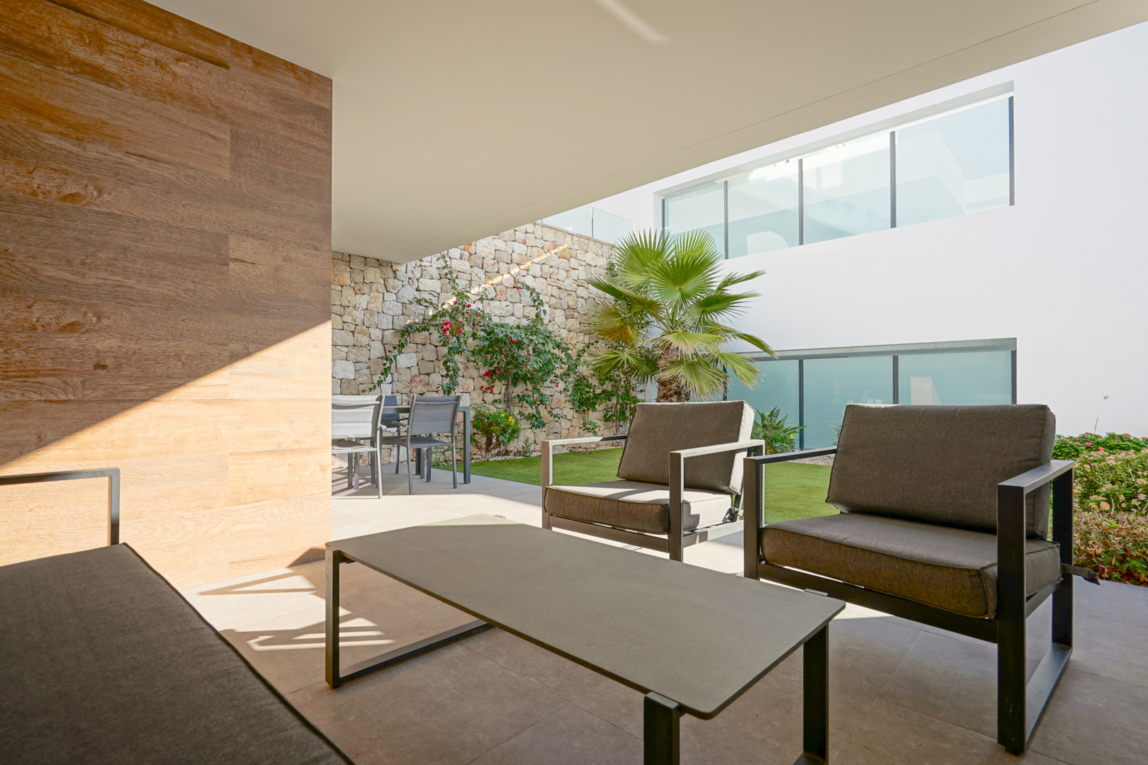 Covered terrace featuring a modern lounge area, dining set, and a private garden bordered by a natural stone wall.