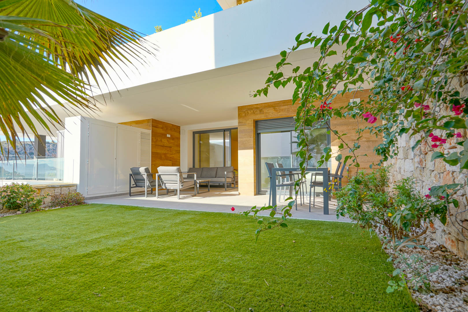Covered terrace featuring modern outdoor seating and dining areas, adjacent to a private garden with artificial lawn and a natural stone wall.