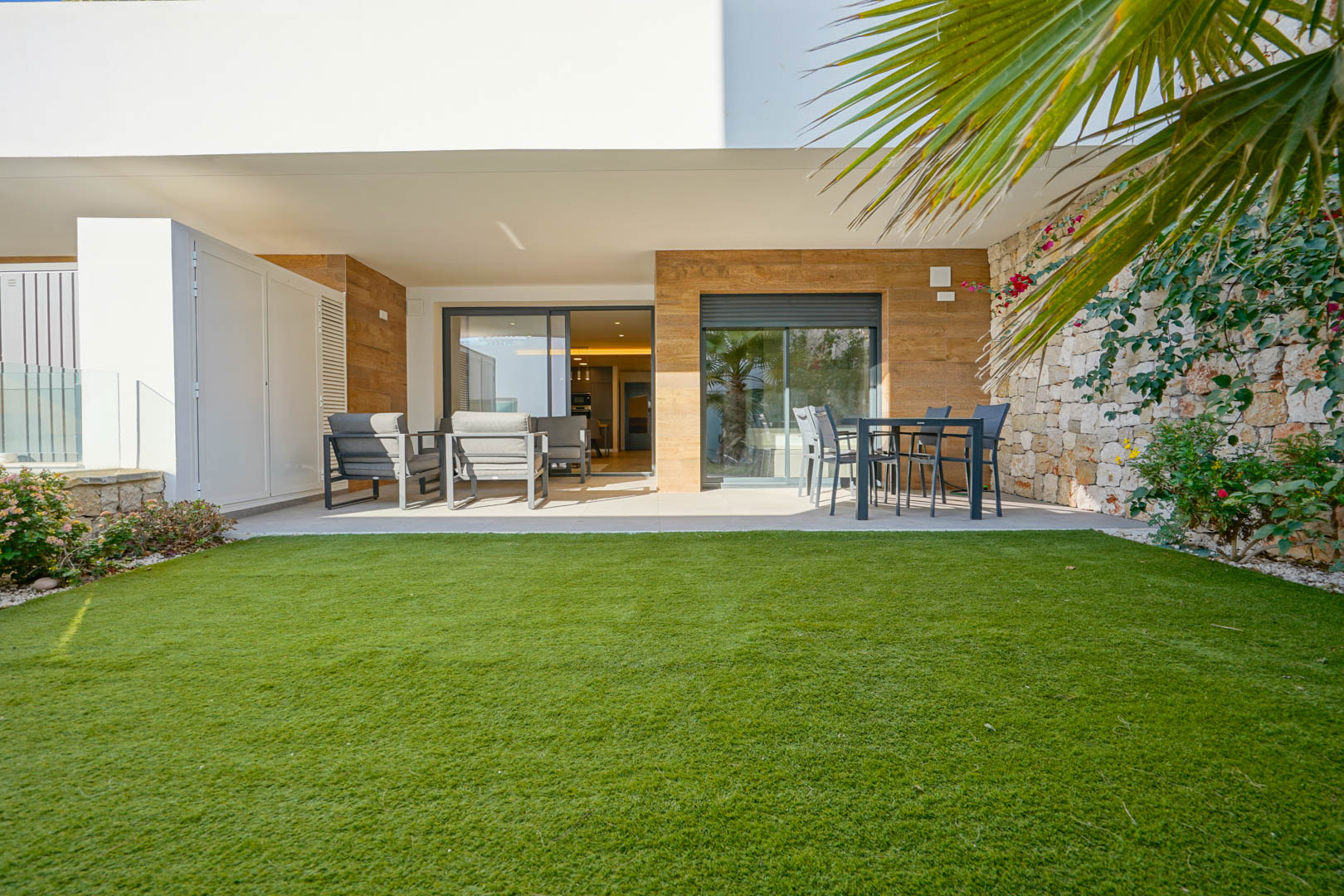 Covered terrace featuring outdoor lounge and dining areas, leading to a private garden with lawn and a characteristic natural stone boundary wall.