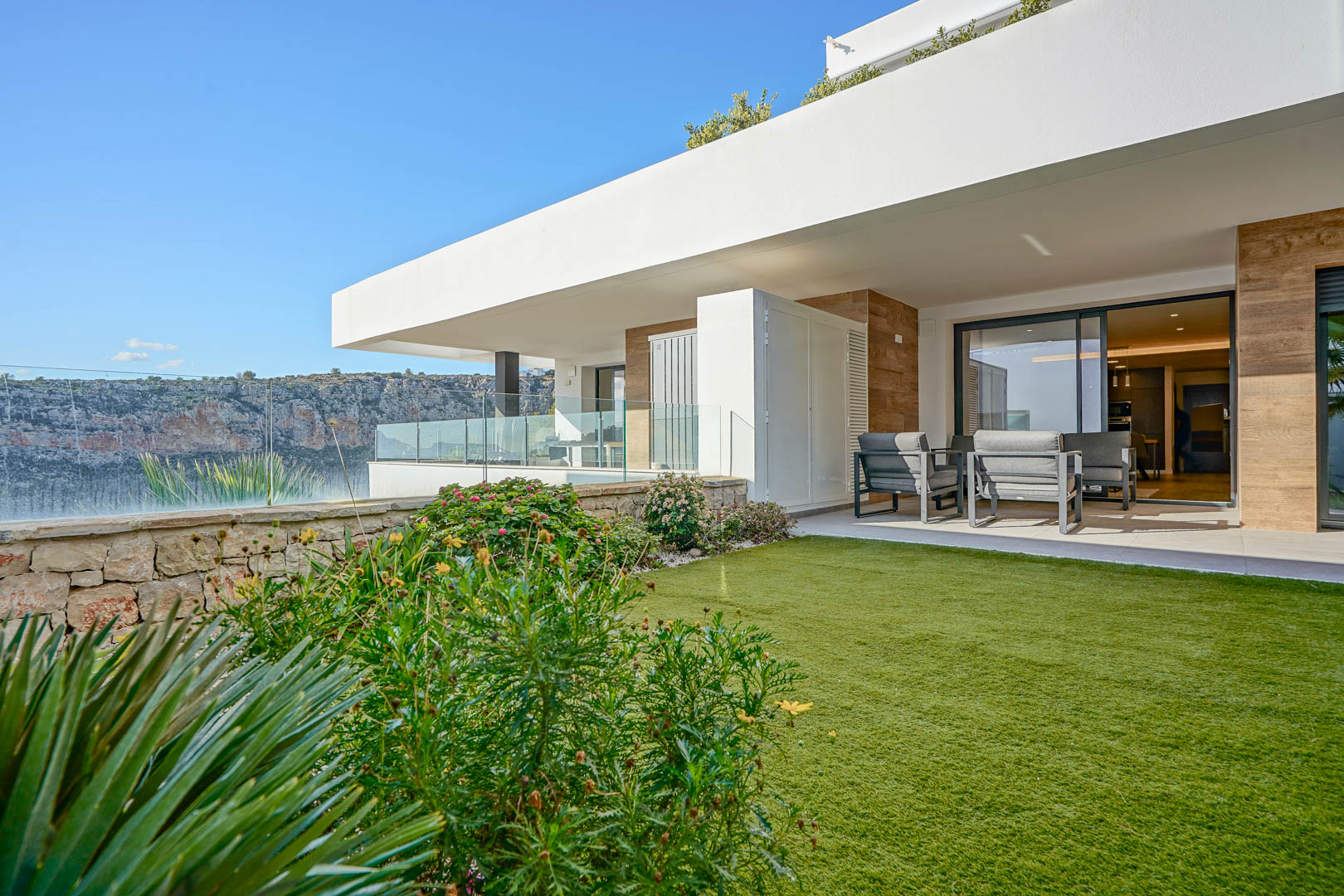 Covered terrace with modern seating and a private lawn area, featuring a natural stone wall and open views of the surrounding cliffs.