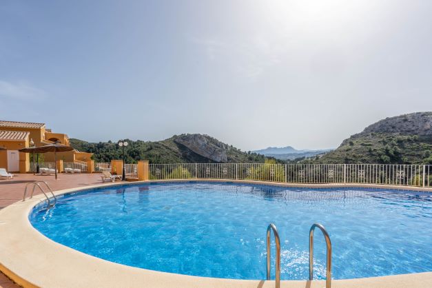Curved communal swimming pool at Monte Cala Gardens, featuring a spacious sun deck with loungers and panoramic views of the surrounding natural park and mountains.