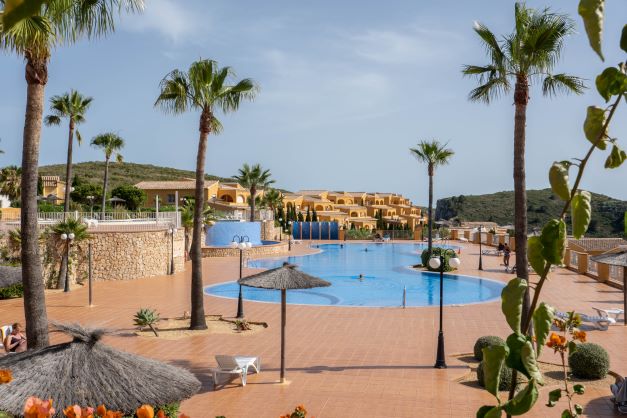 Large communal swimming pool with blue tiling, surrounded by a spacious terracotta terrace, palm trees, and natural stone walls in Monte Cala Gardens.