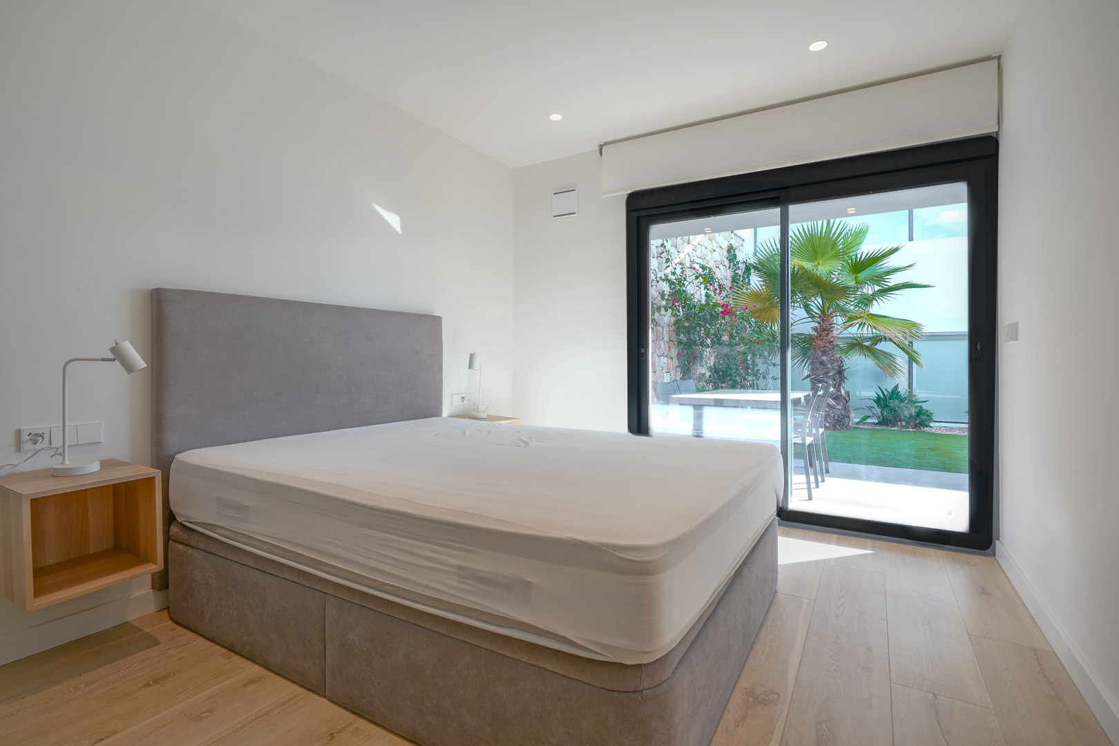 Master bedroom featuring direct garden access through sliding glass doors, light wood flooring, and a contemporary grey upholstered bed frame.