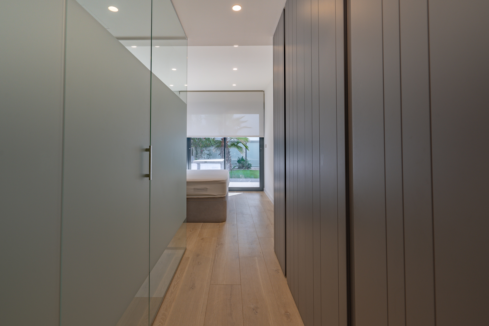 Master bedroom hallway featuring sleek grey built-in wardrobes, a glass partition, and direct views toward the private garden through large windows.