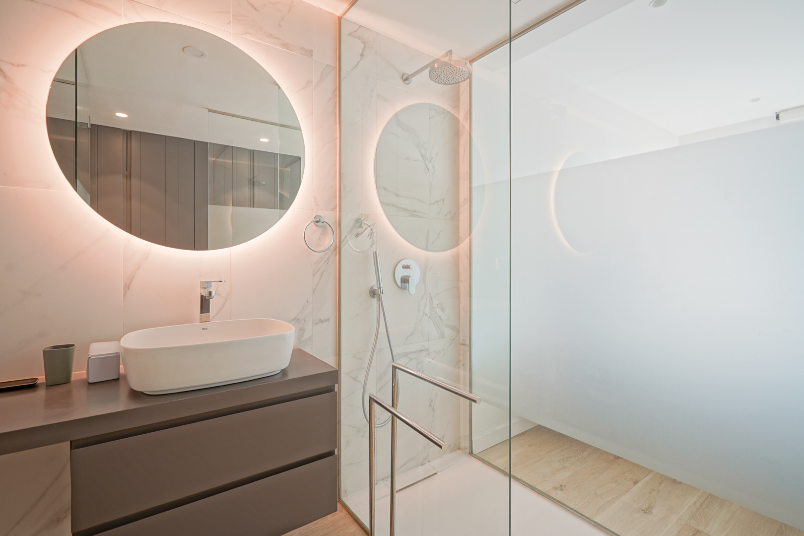 Modern bathroom featuring a vessel sink, backlit circular mirror, and a walk-in shower with glass partition and marble-effect wall tiling.