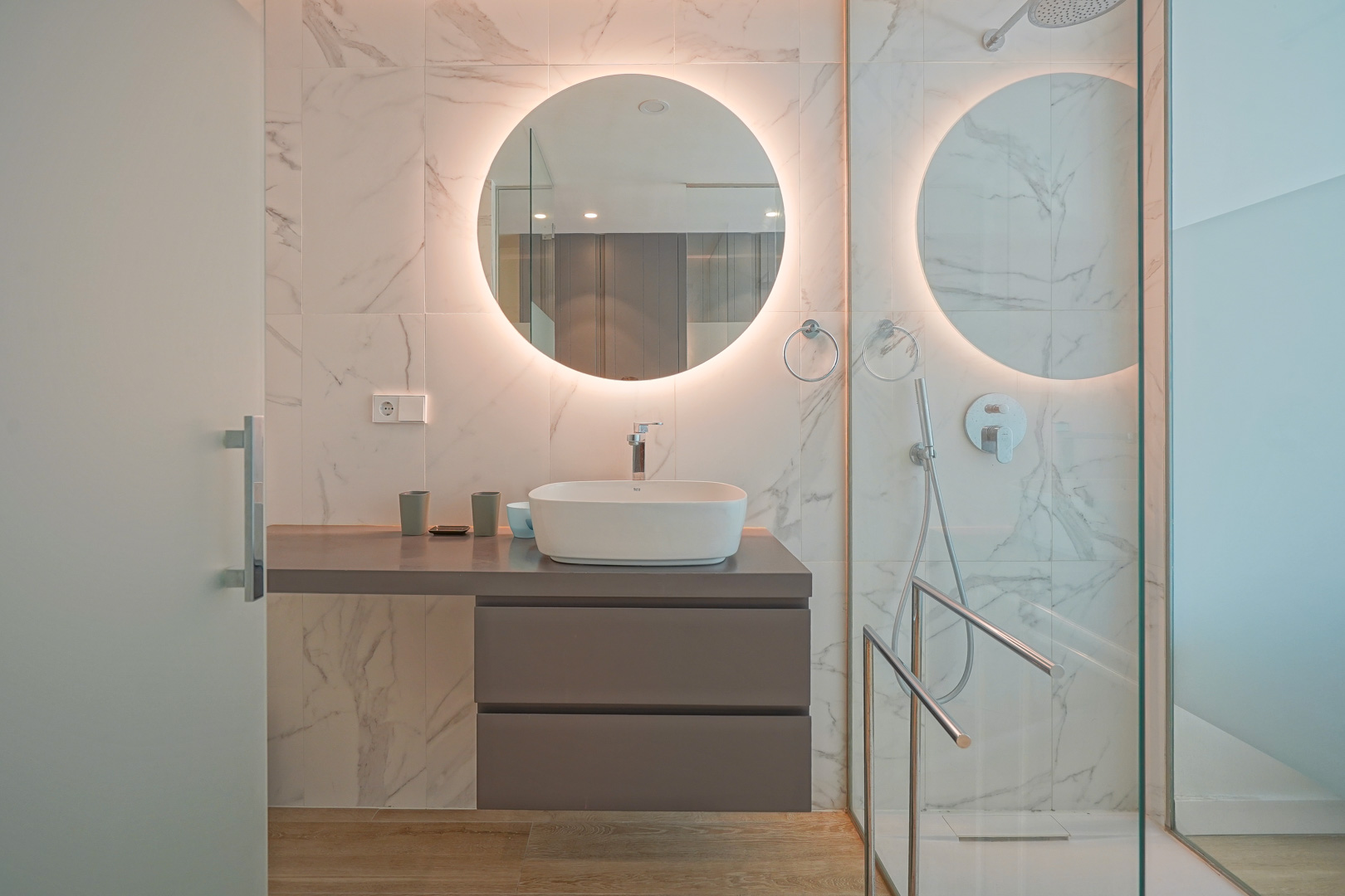 Modern bathroom featuring a vessel sink, backlit circular mirror, and marble-effect wall tiling with a glass walk-in shower.