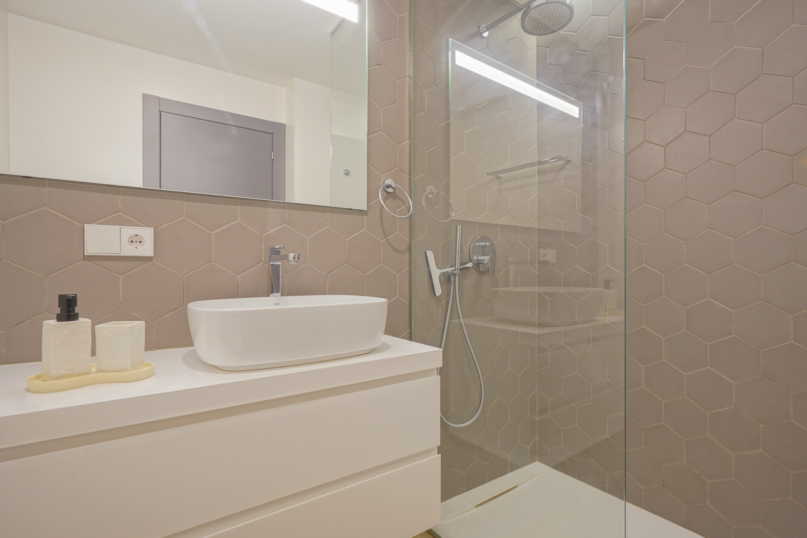 Modern bathroom featuring taupe hexagonal wall tiles, a white vessel sink on a floating vanity, and a walk-in shower with a glass screen.